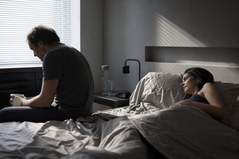 Two people sit apart in a quiet bedroom lit by pale morning light.