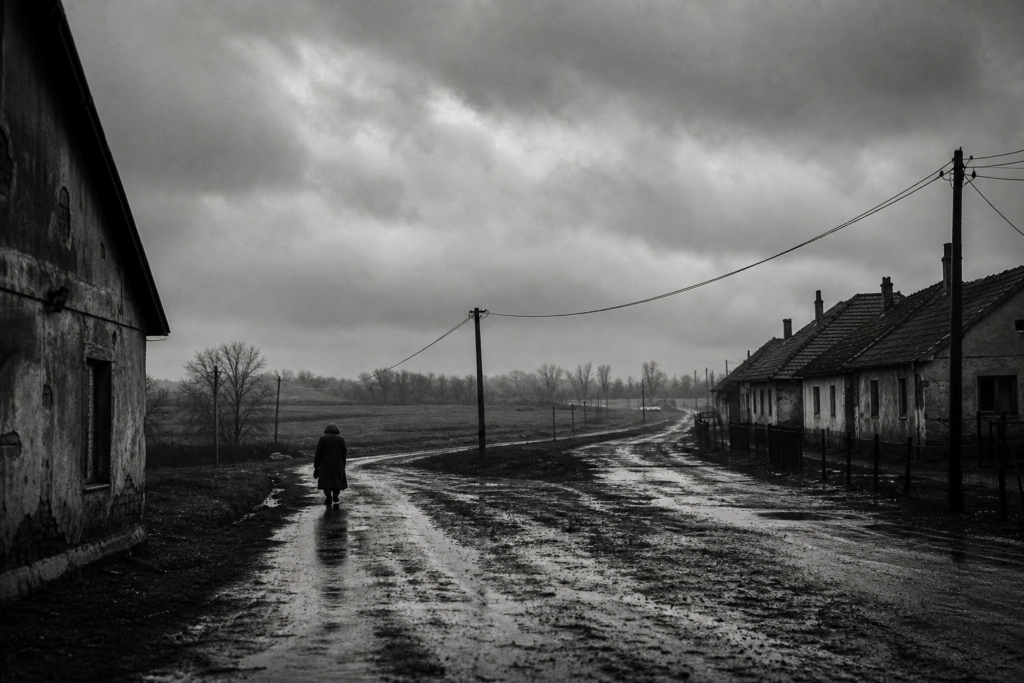 A solitary figure walks along a rain-soaked rural road beneath heavy clouds.